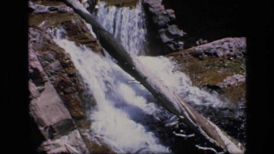 Close 8mm frame of the Montana waterfall, with bright white water, a fallen trunk, exposed rock, and visible film wear.
