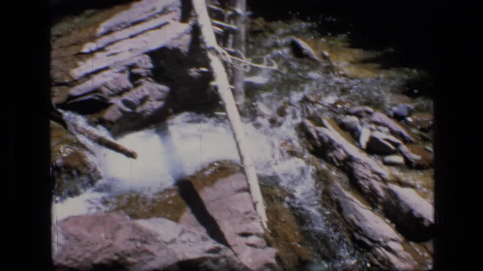 Vintage 8mm footage shows stream water breaking over rocks beside a long pale log in Montana wilderness.