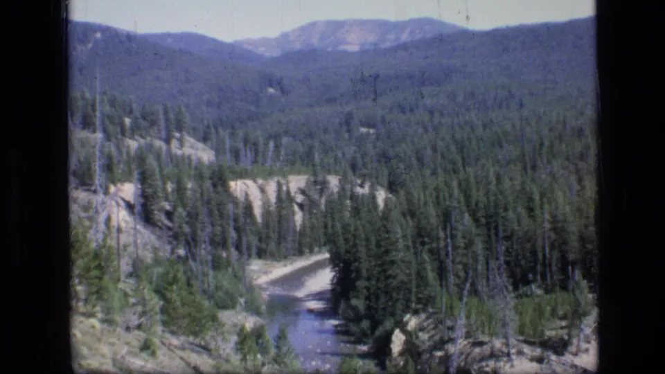 Wide 8mm frame of Scapegoat Wilderness showing a small river below forested slopes and distant Montana mountain ridges.