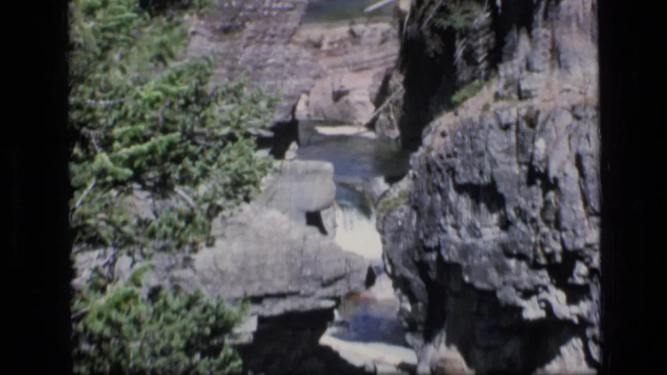 1973 8mm Montana wilderness frame showing a rocky gorge, evergreen trees, and shallow water leading toward a lagoon.