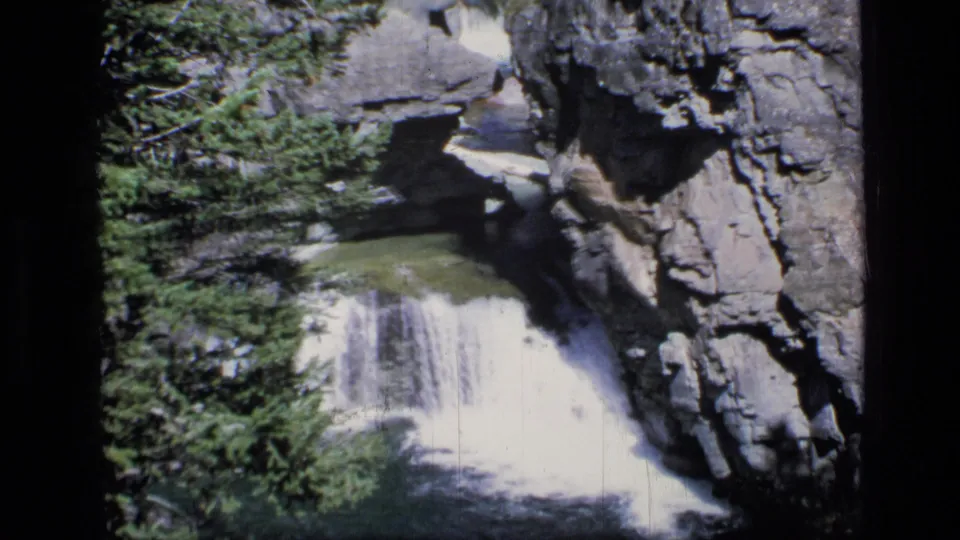 1973 8mm Scapegoat Wilderness view of white water falling into a green canyon pool beside evergreen trees and rock walls.