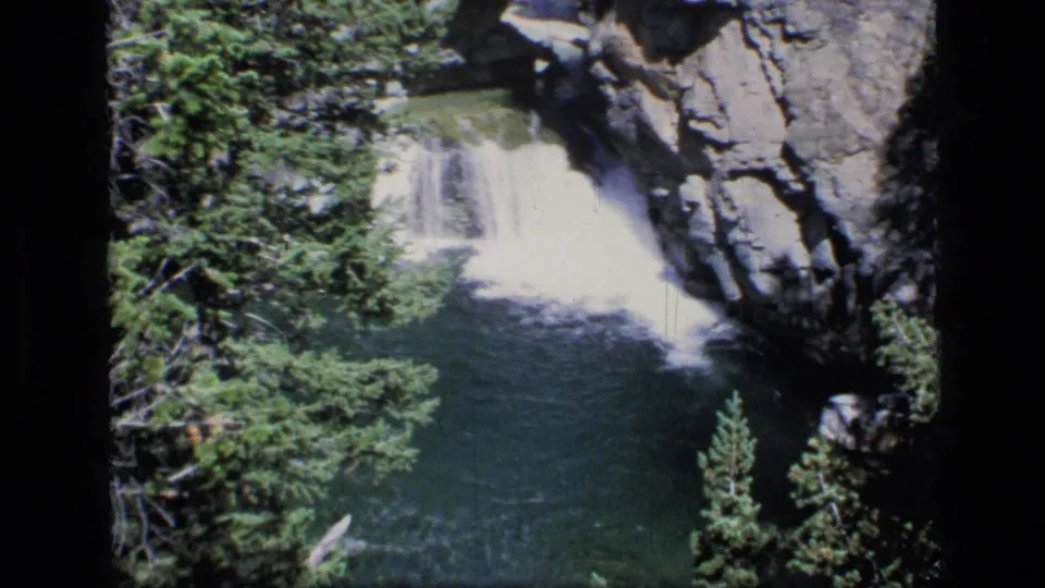 1973 8mm Montana wilderness frame with a bright waterfall, green lagoon, evergreen trees, and dark canyon rock.