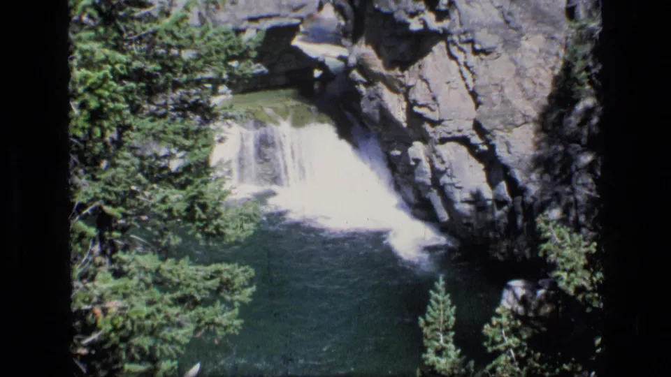 1973 8mm Scapegoat Wilderness view of a waterfall entering a shaded green lagoon below rugged cliff walls.