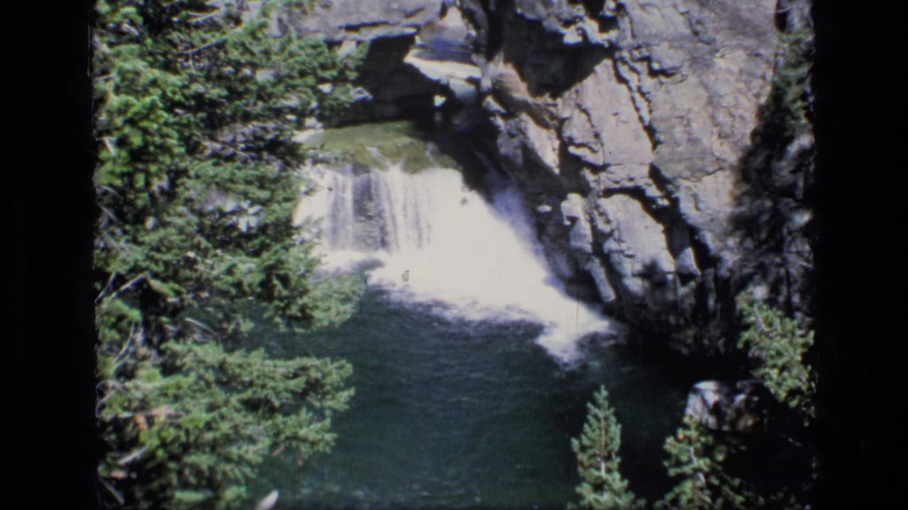 Waterfall drops into a blue-green pool between evergreens and rocky canyon walls.