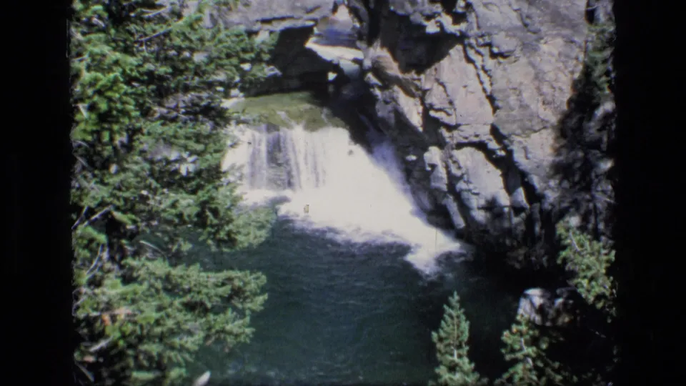 1973 8mm view of a waterfall flowing into a blue-green pool in Montana wilderness, framed by evergreens and rock walls.