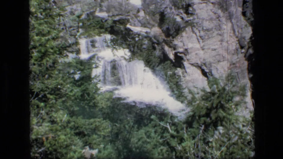 Waterfall in Scapegoat Wilderness appears behind dense evergreen trees with pale rock cliffs rising beside the cascade.