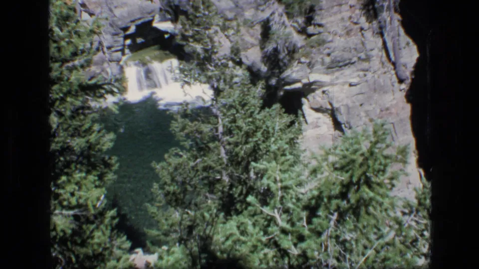 Handheld 8mm frame shows the waterfall feeding a shaded pool below evergreen trees and layered rock ledges.