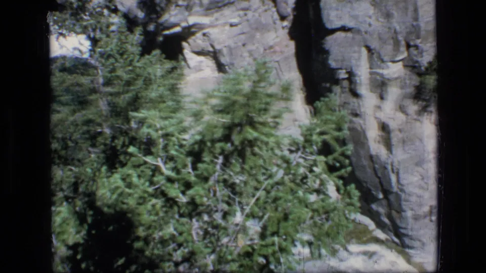 Late frame from the 1973 wilderness clip shows handheld motion past evergreens, with the waterfall and cliff partly obscured.