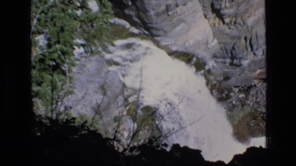 1973 8mm Montana wilderness frame showing a bright waterfall dropping along a gray rock face with trees and brush nearby.
