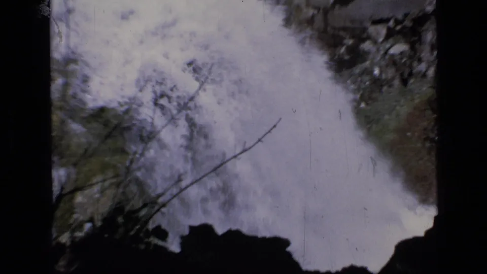 1973 8mm Scapegoat Wilderness frame showing foaming water below the falls, with rocks, brush, and a thin branch crossing the view.