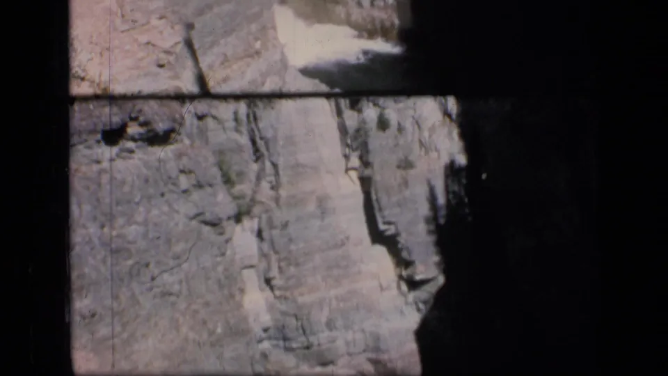1973 Montana wilderness frame showing a steep canyon face and a pale distant watercourse or waterfall against layered rock.