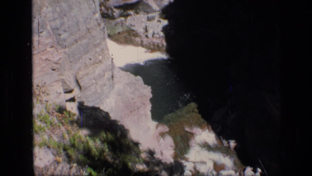 1973 8mm view of Scapegoat Wilderness terrain, looking down into a rocky canyon with shadowed water between steep stone walls.