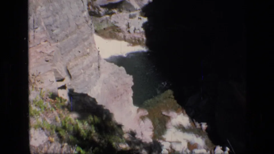 1973 8mm view of Scapegoat Wilderness terrain, looking down into a rocky canyon with shadowed water between steep stone walls.