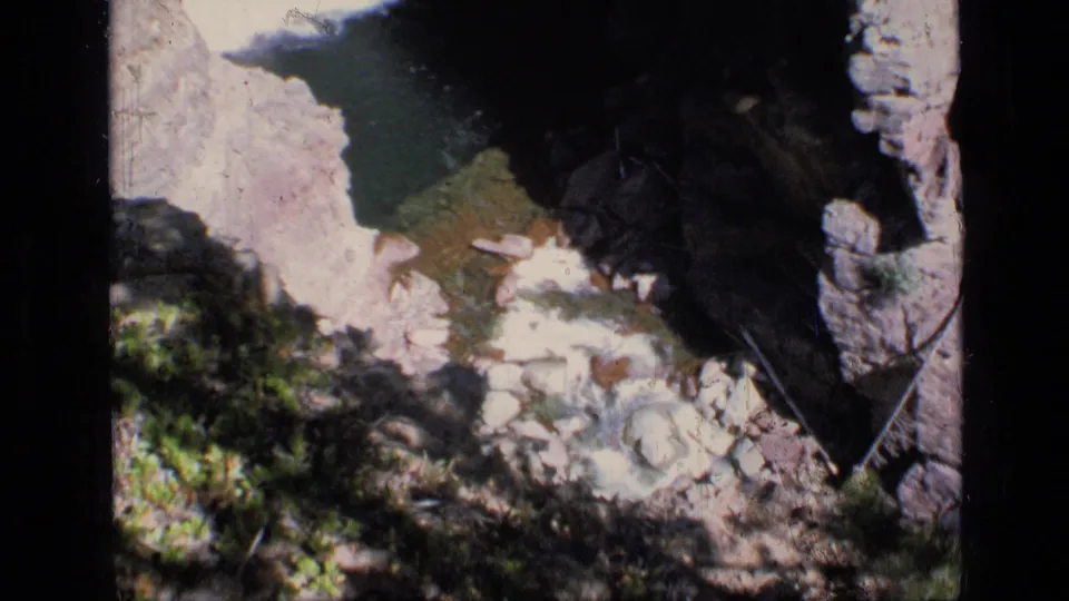 The 1973 home-movie frame looks down toward a rocky Montana canyon floor with water, pale stones, dark shade, and low green vegetation.