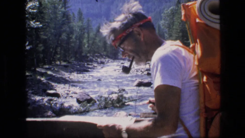 Adult man in 1973 Scapegoat Wilderness footage studies a map while smoking a pipe beside a rushing river and forested bank.