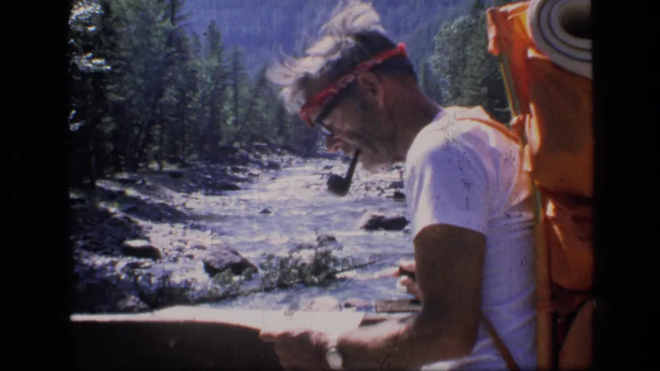 The hiker remains beside the river in the supplied Montana wilderness scene, holding a map with his orange pack visible.