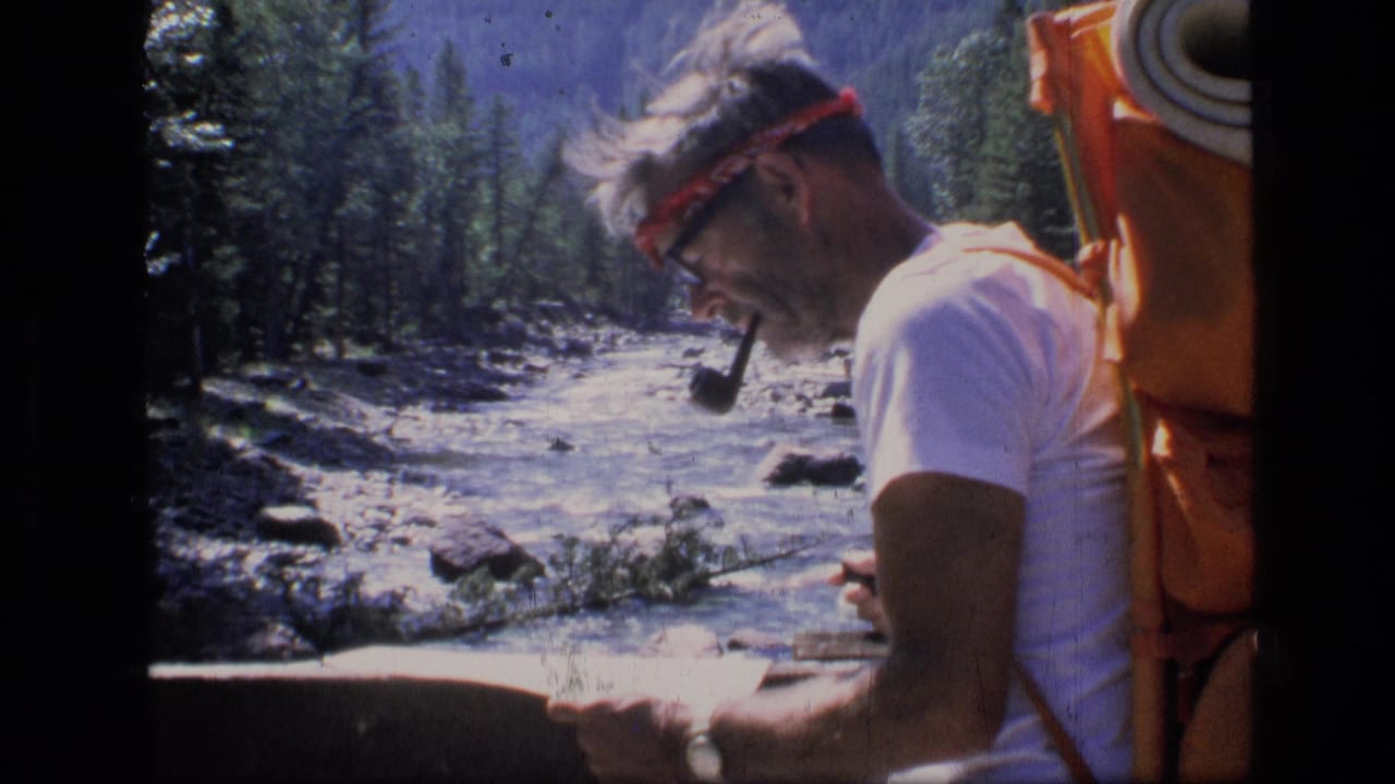 Man with pipe and red headband looks down at a map beside a rocky river.