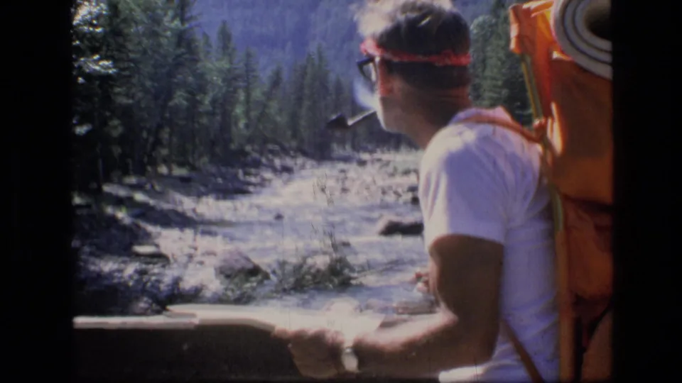 A backpacker in Scapegoat Wilderness, Montana, sits at the river edge with a pipe and red headband, facing the trees and flowing water.