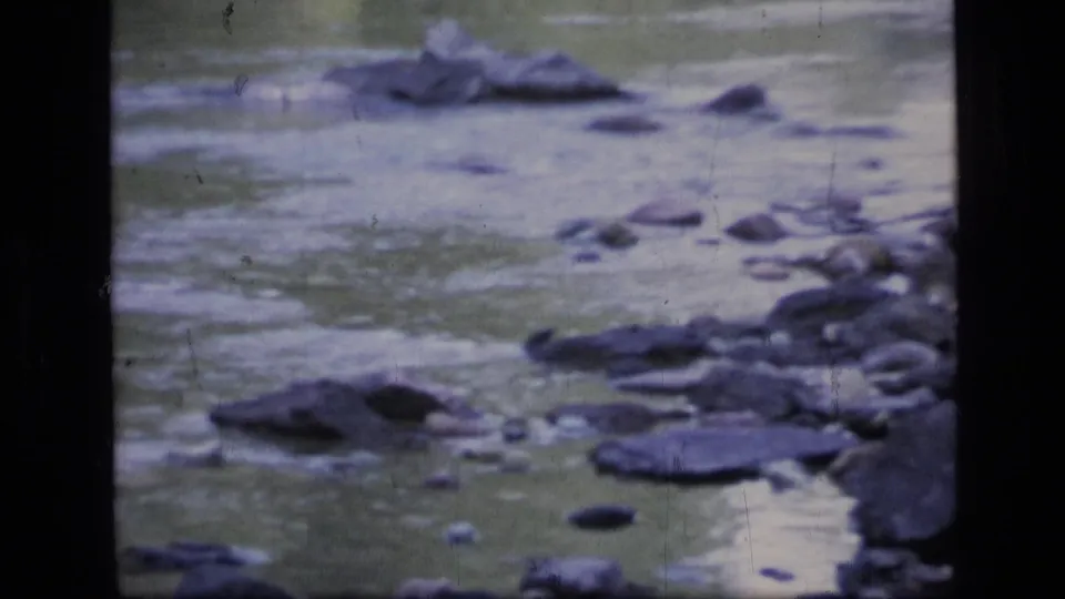 A close river cutaway shows shallow moving water and scattered rocks in the Montana wilderness, rendered with soft 8mm color and grain.