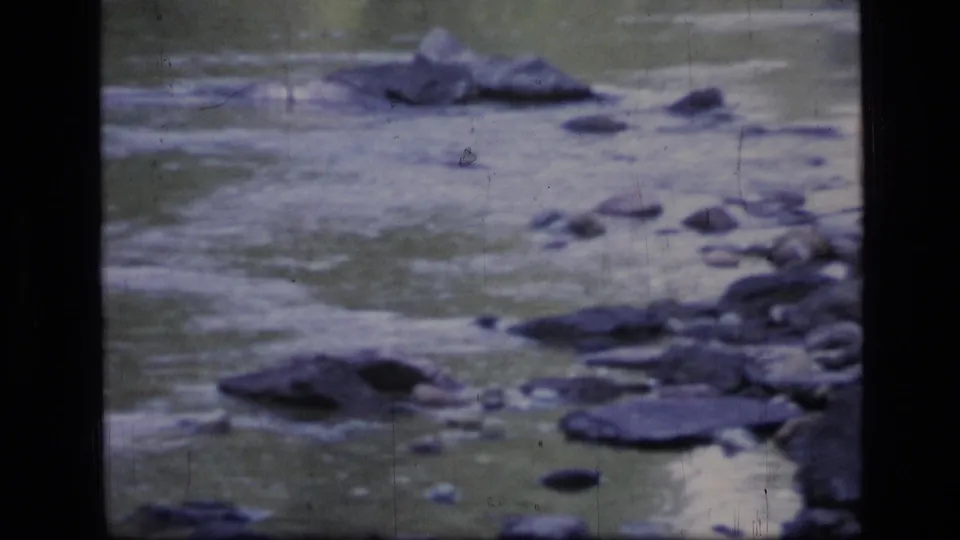 Archival 8mm view of a Scapegoat Wilderness stream, with rounded stones, muted water tones, and a dark film gate edge.