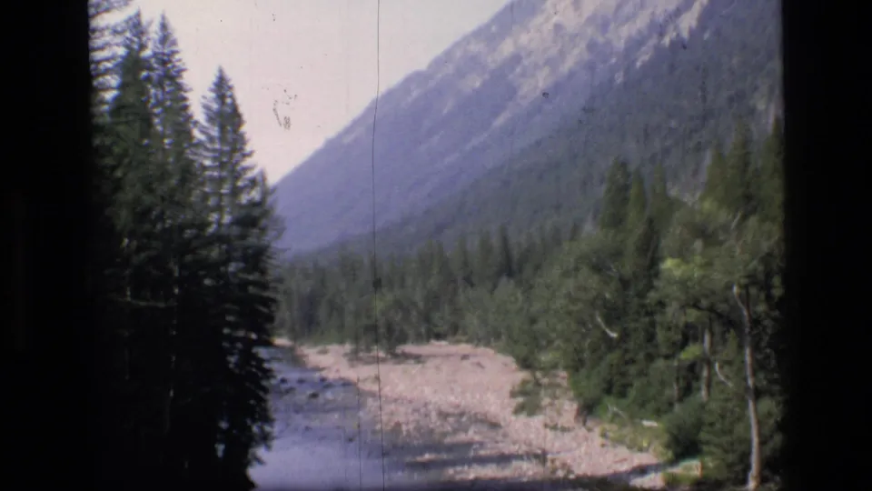 1973 8mm frame of a rocky streambed and dense evergreens beneath steep Scapegoat Wilderness mountain slopes.