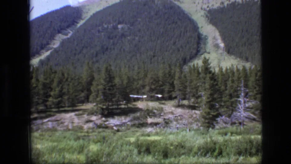 Wide 1973 Scapegoat Wilderness frame showing a distant structure in a pine clearing below forested hills with open lanes.