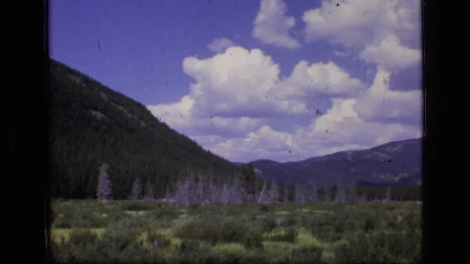 1973 8mm view of Scapegoat Wilderness meadow, forest, and distant mountains under large white clouds.