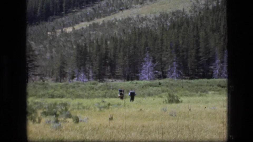 Final 8mm frame shows two distant hikers in a Montana meadow with evergreen forest and mountain slope behind them.