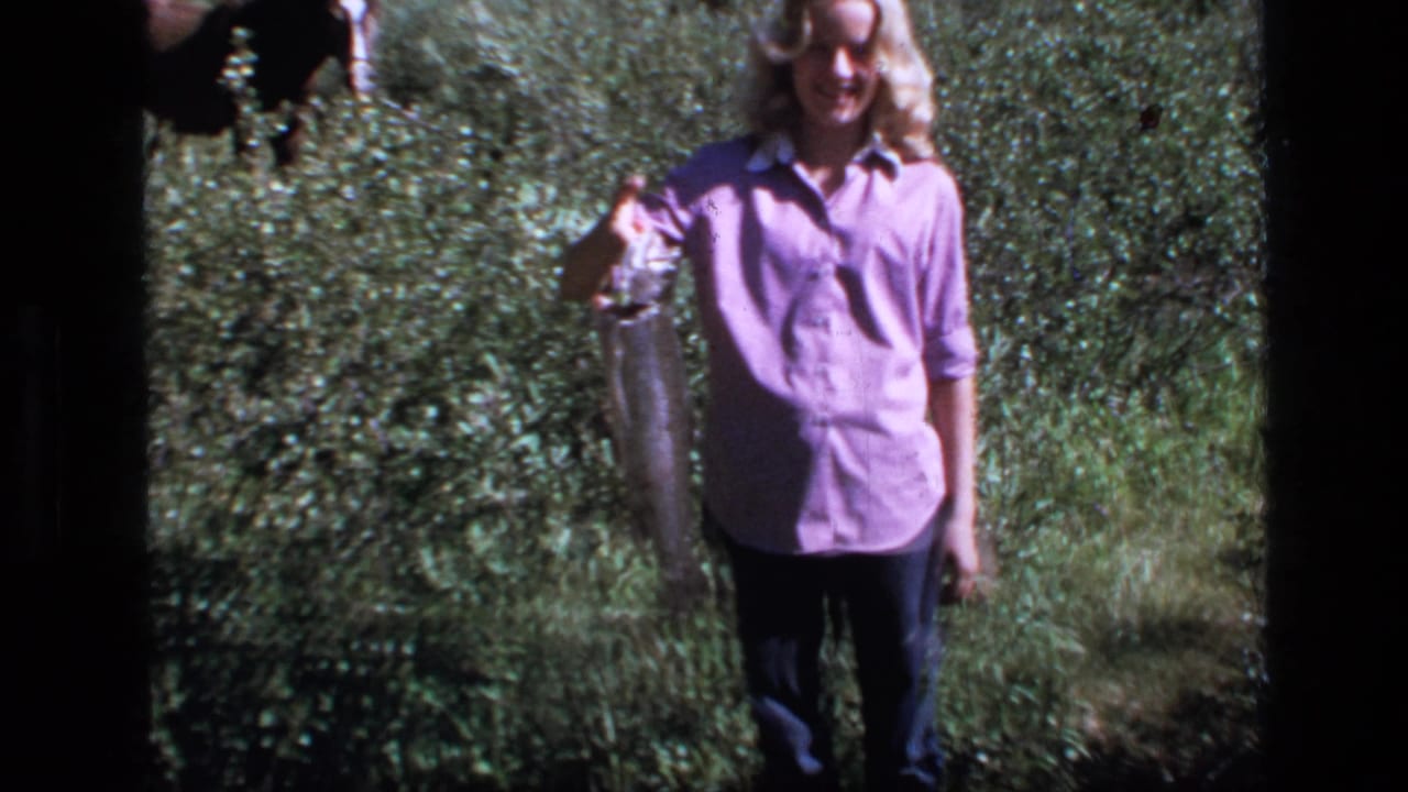 1973 Montana home-movie frame showing a young woman holding a large fish in grassy wilderness.