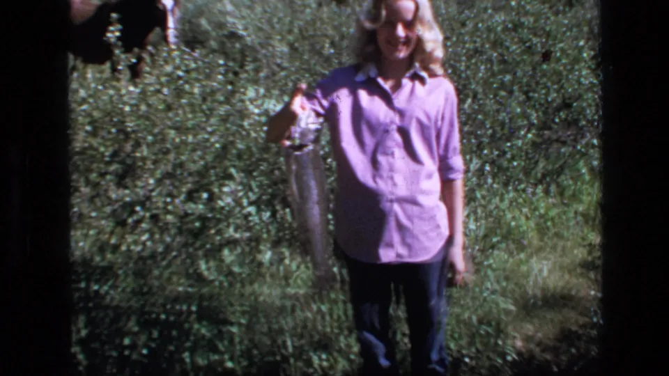 1973 Montana home-movie frame showing a young woman holding a large fish in grassy wilderness.