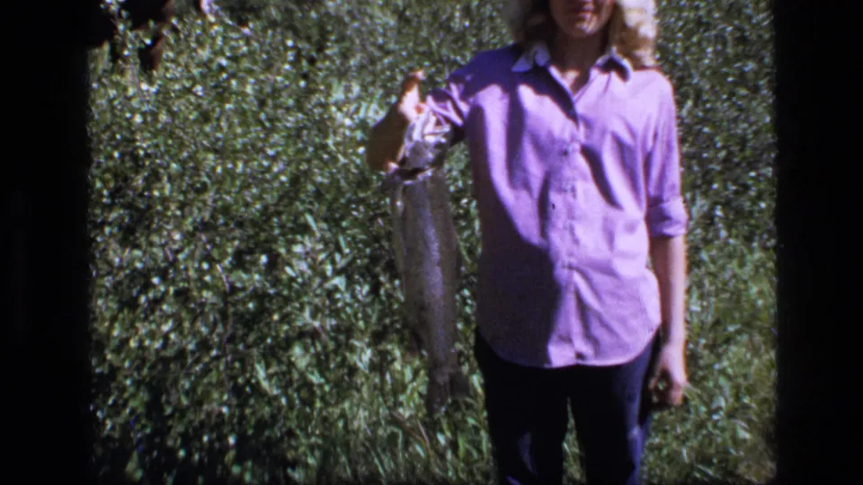A closer 8mm frame of a young woman in a purple shirt holding a large fish outdoors in Montana.