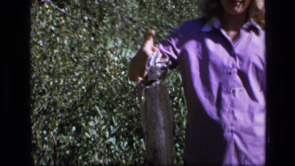 Close handheld 8mm view of the young woman presenting the fish against tall grass and brush.