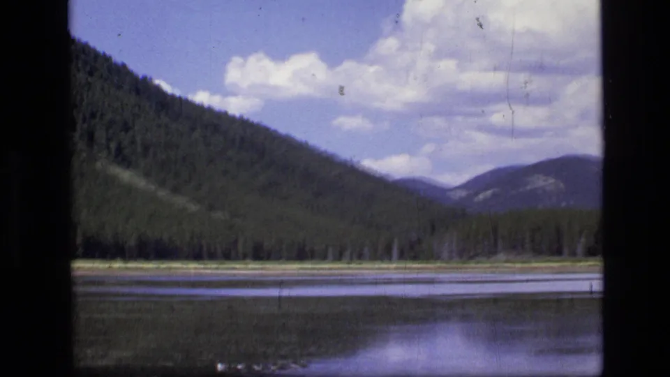 1973 Montana wilderness frame with a still lake, dark evergreen shoreline, mountain slopes, and bright clouds above.