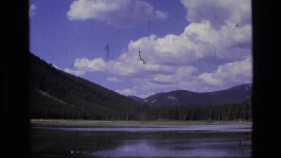 1973 8mm Scapegoat Wilderness view across a mountain lake, with small ducks low in frame and clouds over forested ridges.