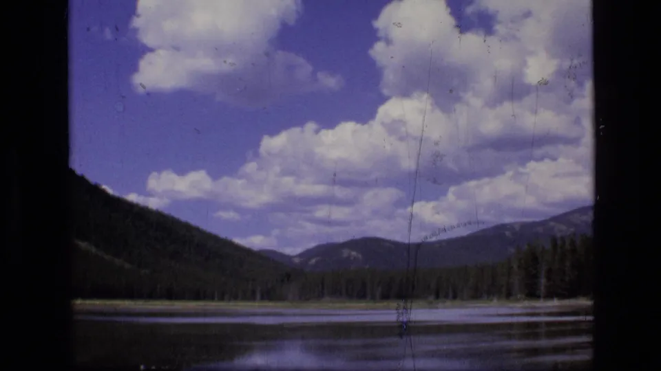 1973 Montana wilderness frame with a quiet lake, evergreen shore, distant mountain ridges, and large white clouds in a blue sky.