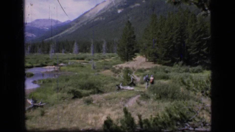 1973 8mm view of Scapegoat Wilderness, Montana, with distant hikers on a meadow trail near a stream, pines, and mountain slopes.