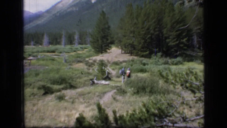 1973 Montana wilderness frame showing backpacked hikers on a curving meadow trail with pine forest, stream, fallen logs, and mountains beyond.
