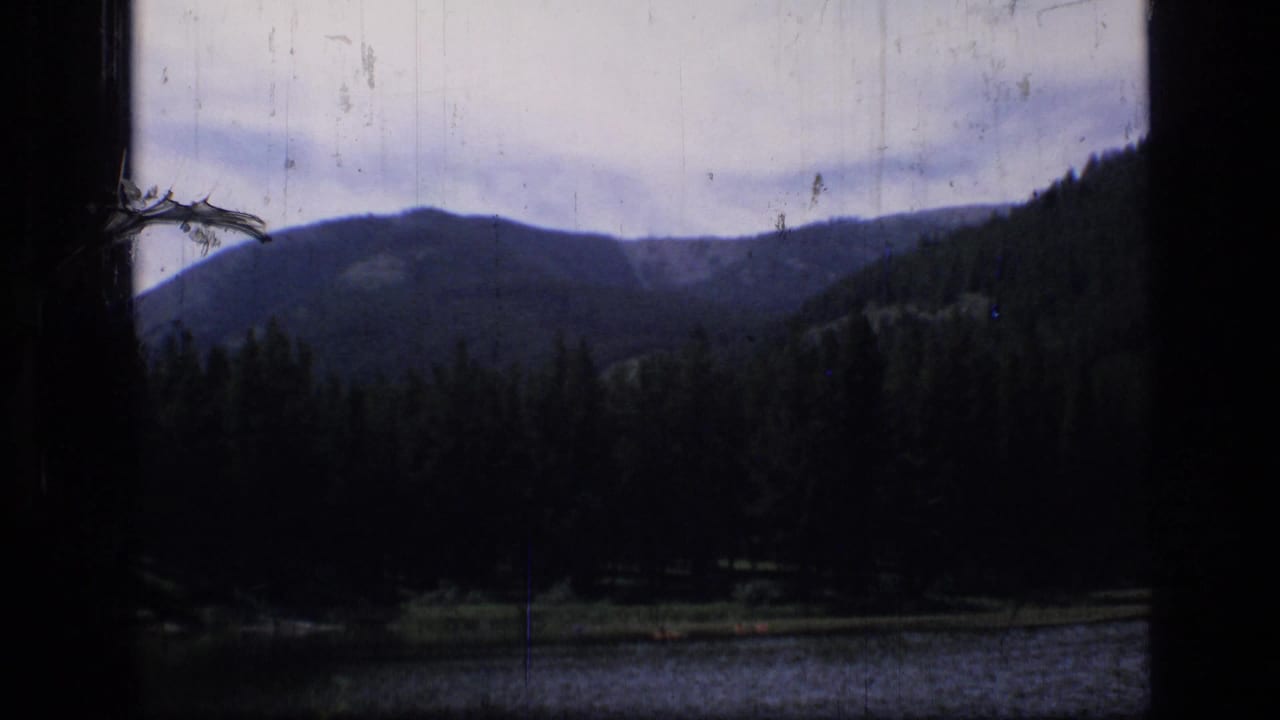 1973 8mm view in Scapegoat Wilderness, Montana, showing a shaded mountain lake, evergreen trees, and distant ridges framed by dark film edges.