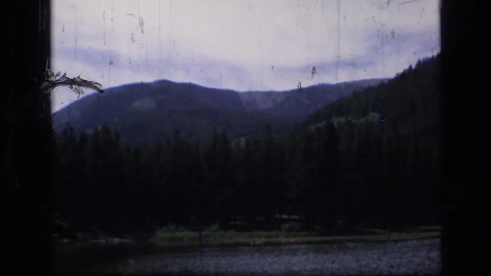 1973 8mm view in Scapegoat Wilderness, Montana, showing a shaded mountain lake, evergreen trees, and distant ridges framed by dark film edges.