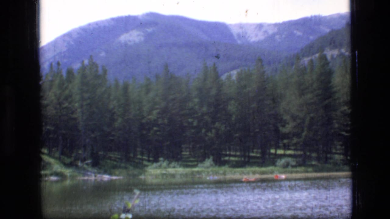 Lake shoreline with evergreen trees, mountains, and small red boats near the water.