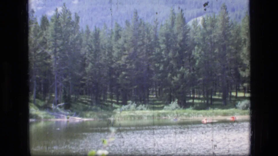 1973 8mm Scapegoat Wilderness view of a forested shoreline, mountain lake water, faint ridges, and a small red boat in the lower frame.