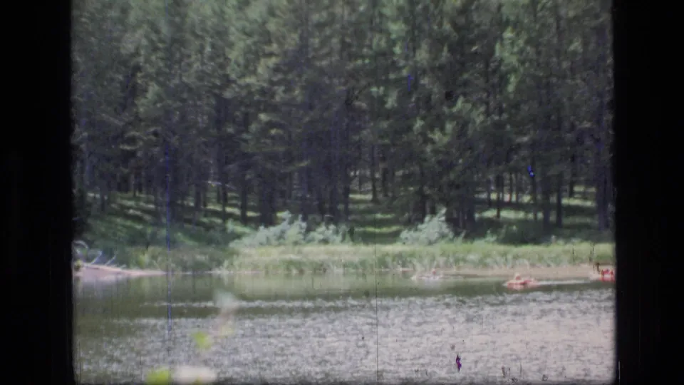 1973 Montana lake scene showing dense evergreens, grassy shoreline, calm water, and small boats or figures near the right edge.