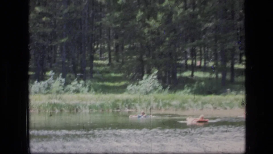 Distant people in the lake appear near a red raft against the evergreen shoreline in this 1973 Montana home-movie frame.