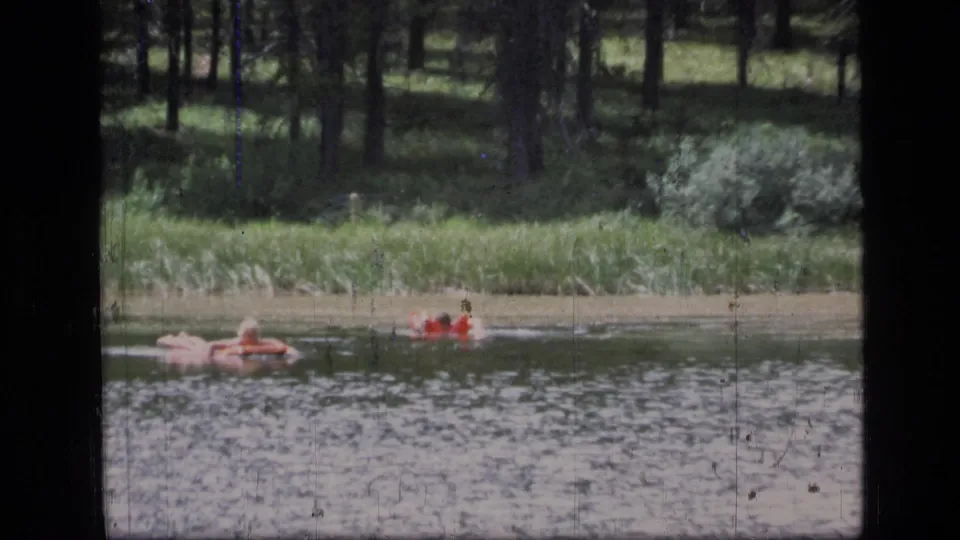 A closer 8mm frame shows people floating near a red raft on a forest lake in Scapegoat Wilderness, Montana.