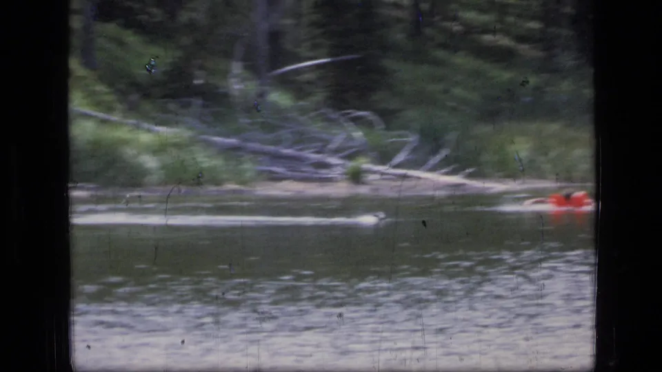 The red raft drifts at the edge of the frame while rippled water, forest, and fallen branches fill the Montana lake scene.