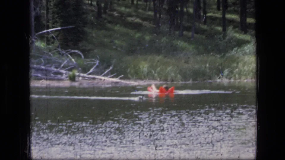 In 1973 Scapegoat Wilderness footage, a red raft appears on a lake with trees and grass along the far bank.