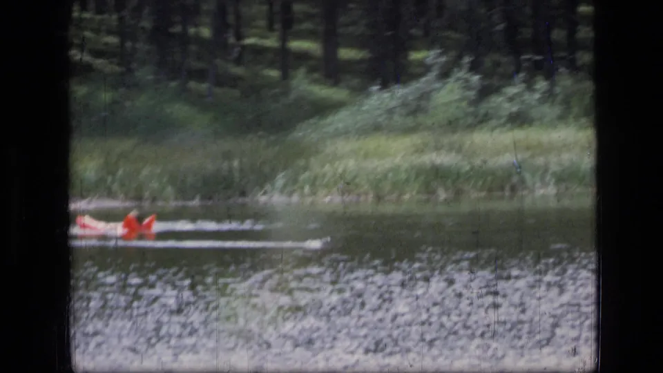 The red raft continues across the Montana lake, leaving a pale wake in front of a green forested shoreline.