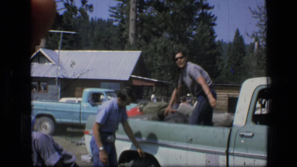 Men handle gear around a light-colored pickup near a cabin and trees in 1973 Montana home-movie footage.