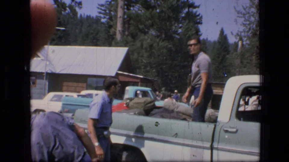 A man stands by a loaded pickup while others work nearby, with a cabin and Montana forest visible behind.