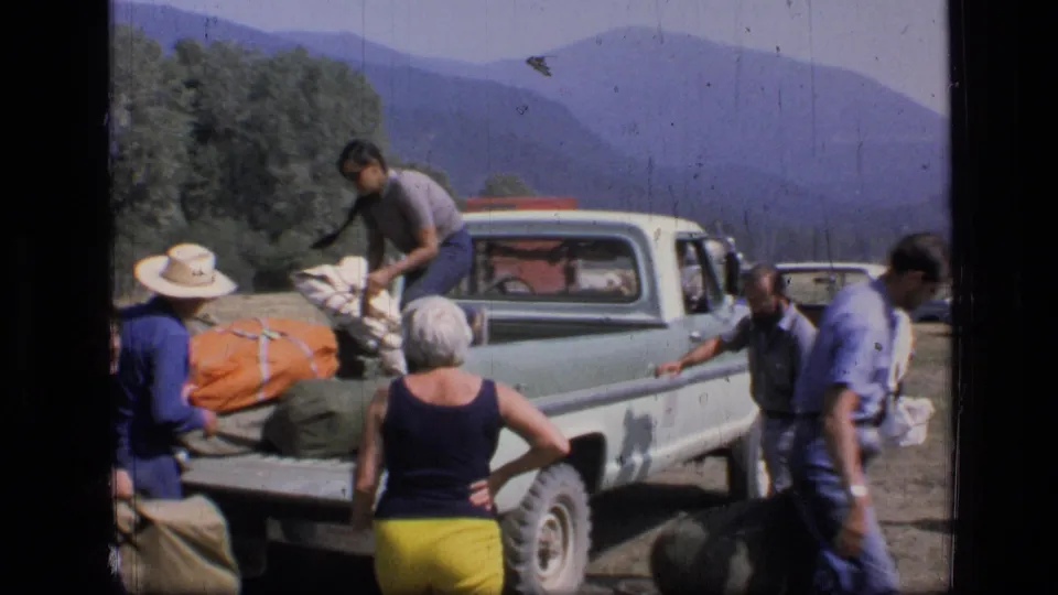 Home-movie frame of adults unloading orange and green camping bags from a vintage pickup in a Montana mountain setting.